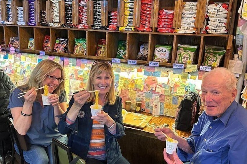 Three people eating from paper cups in a store with shelves of snacks and vegan labels. 2