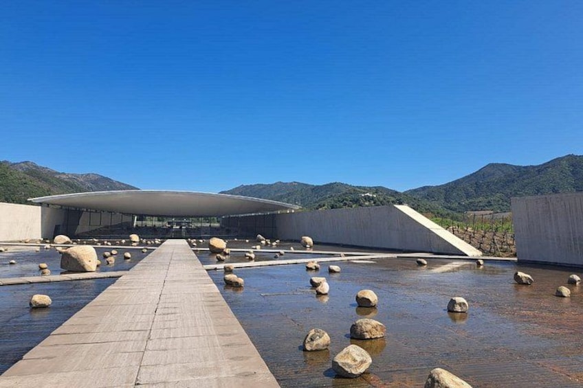 A modern building with a curved roof, a concrete pathway, and scattered rocks in a water-filled area with mountains in the background. 4