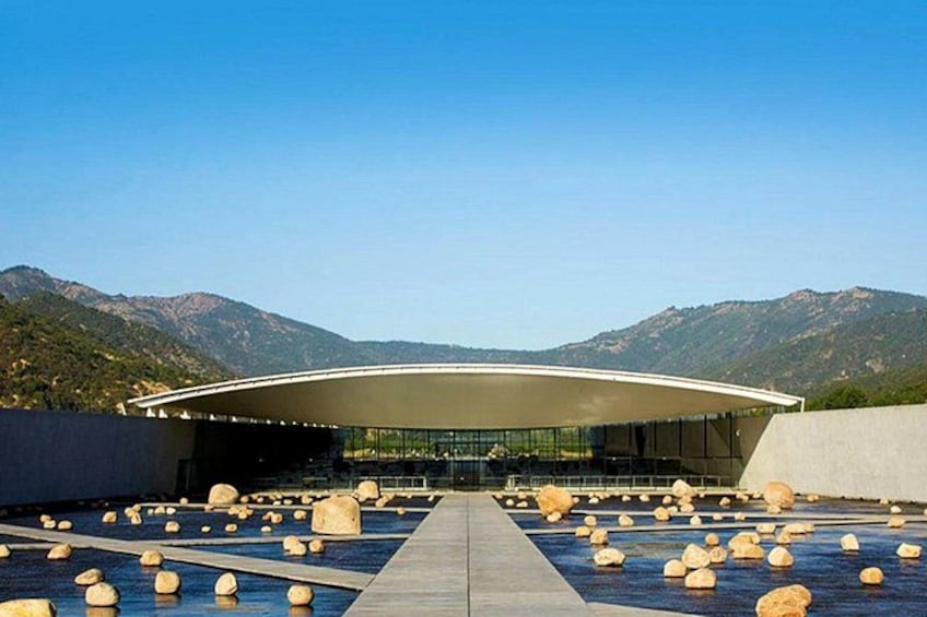 A modern building with a curved roof, surrounded by a pool with large rocks, set against a mountainous backdrop. 1