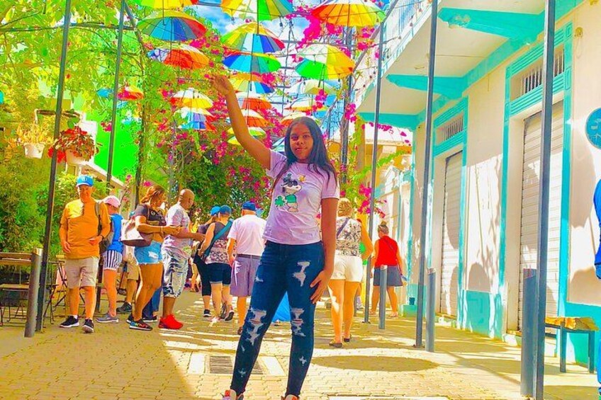 A person standing under a canopy of colorful umbrellas, with buildings and pedestrians in the background. 1