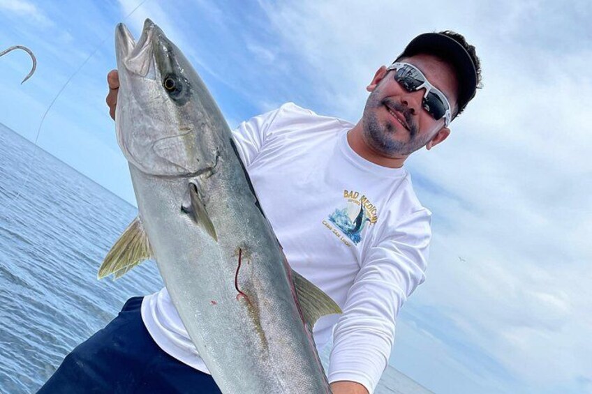 Eine Person hält einen großen Fisch, trägt ein weißes Langarmshirt und steht auf einem Boot, im Hintergrund ist ein klarer Himmel zu sehen. 4