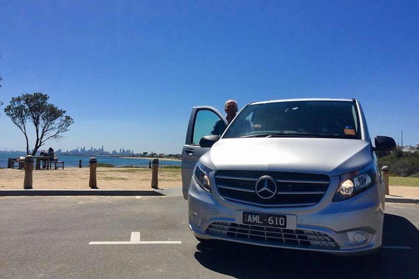 A silver Mercedes-Benz parked on a beachside road with a clear blue sky and a distant city skyline. 1