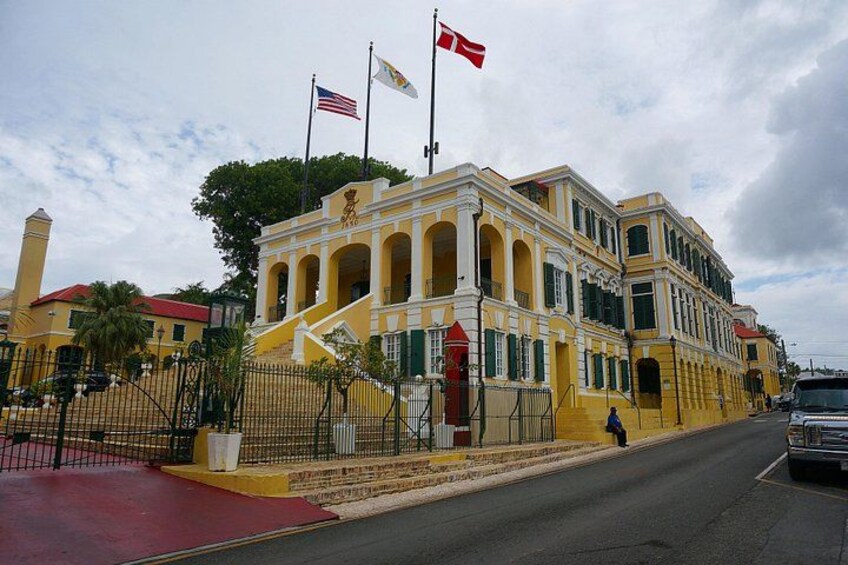 A yellow building with a red roof, a flagpole with flags, and a staircase leading to the entrance. 3