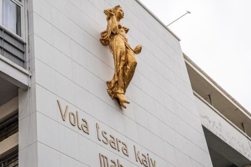 A building facade with a golden statue and the inscription 'Vola tsara kai' on a white wall. 4