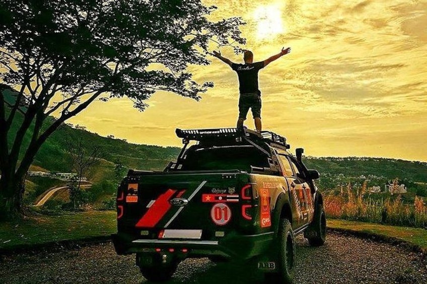 Un camionnette dont on voit l’arrière, avec une personne debout sur la benne, un arbre et un coucher de soleil en arrière-plan. 2