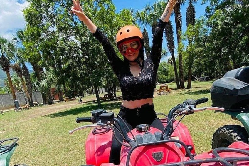 A person with arms raised, wearing a helmet and a black top, standing next to a red ATV in a grassy area with trees and a clear sky. 4