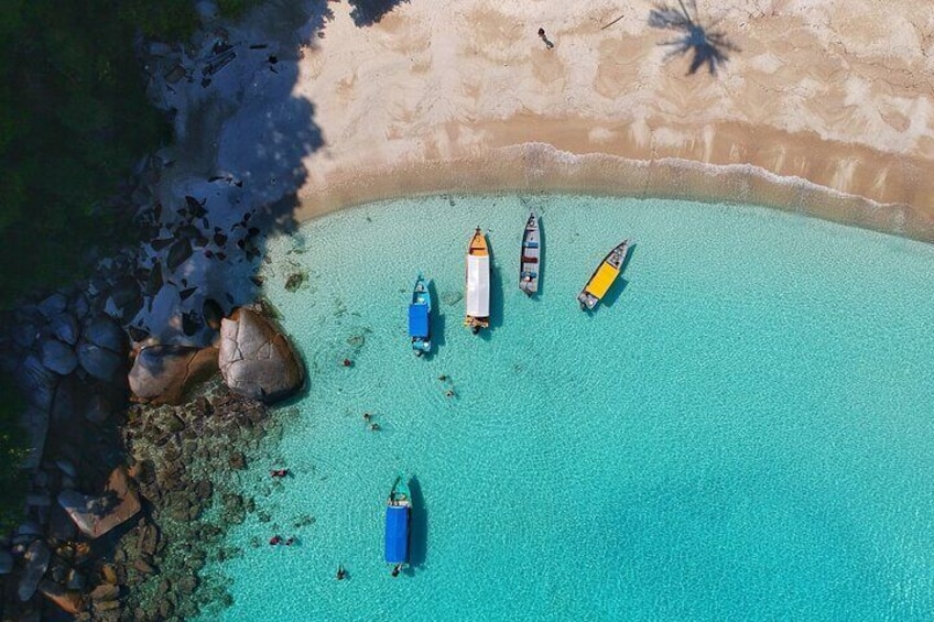 An aerial view of a turquoise beach with several boats anchored near the shore, surrounded by rocks and palm trees. 3