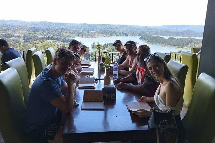 A group of people seated at a table with a scenic view of a lake and hills in the background. 2