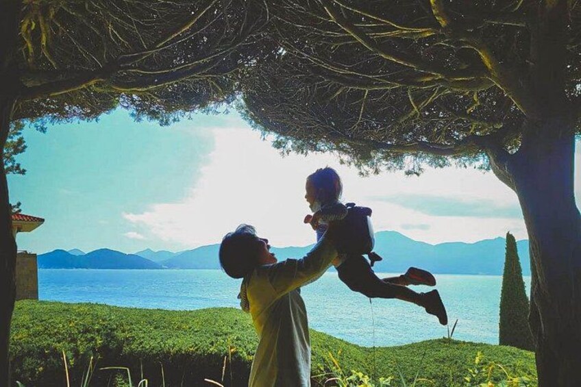 Una persona sosteniendo a un niño bajo un árbol, junto a un lago con montañas al fondo. 2