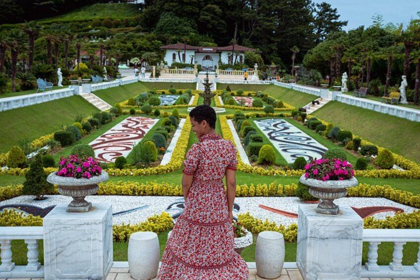 Una mujer con un vestido rojo parada en un jardín con una fuente, césped cuidado y un edificio al fondo. 3