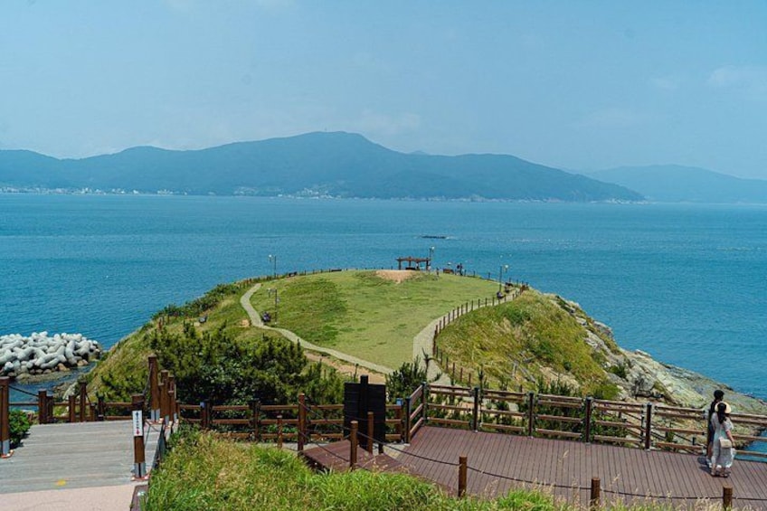 Un paseo marítimo junto a la costa con un área verde, cerca viva de madera y vistas al mar y montañas distantes. 2