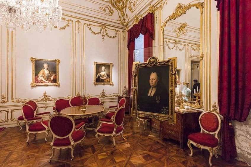 An ornate room with a large portrait, red chairs, and a chandelier. 3