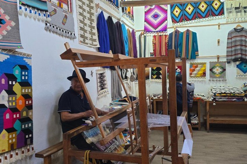 Una persona tejiendo en un telar de madera, con textiles y diseños coloridos exhibidos al fondo. 2