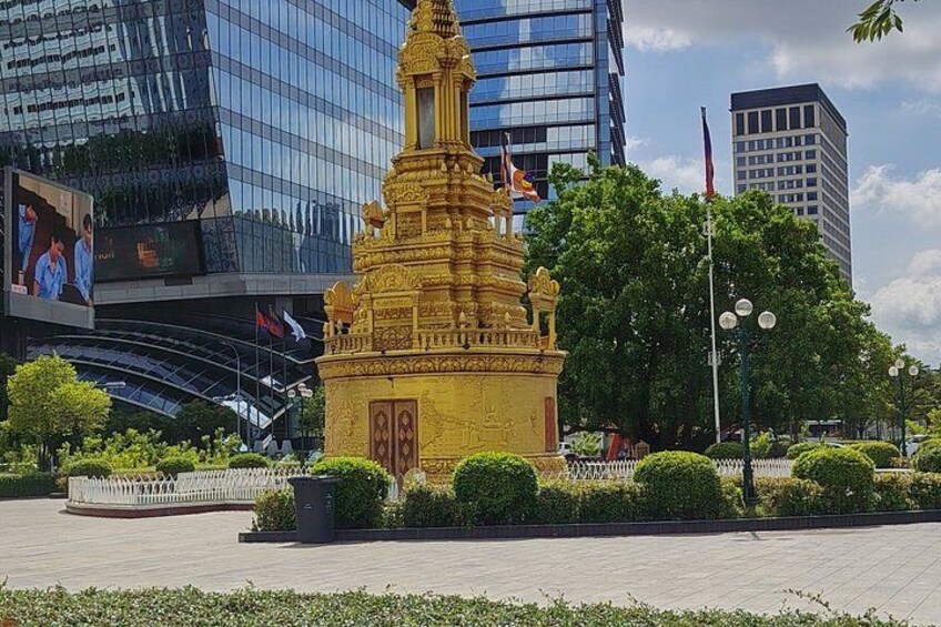 A golden stupa in a city square with modern buildings and trees in the background. 2