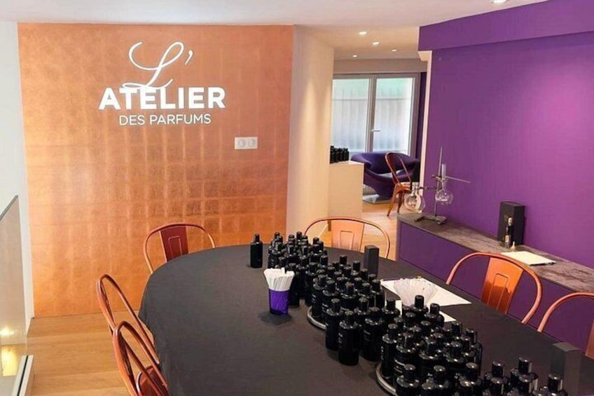 Et moderne interiør med et rundt bord dækket til et møde, en lilla accentvæg og et skilt med teksten 'ATELIER DES PARFUMS'. 1