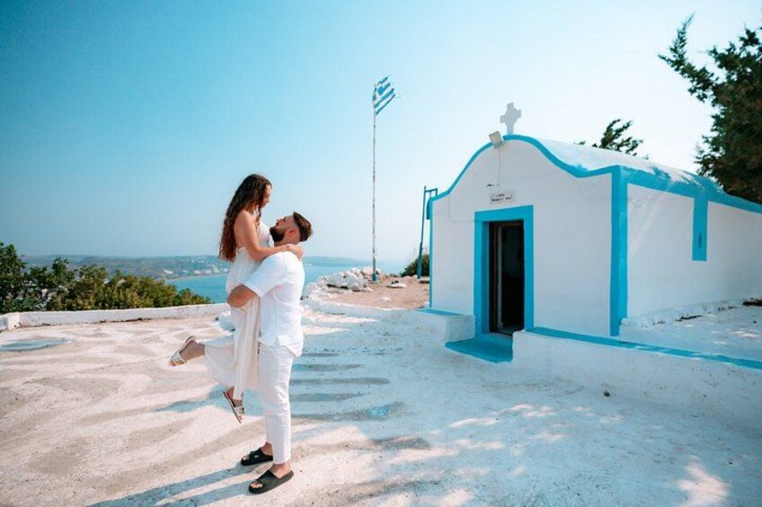Ein Paar umarmt sich am Strand, im Hintergrund sieht man eine kleine Kirche. 1