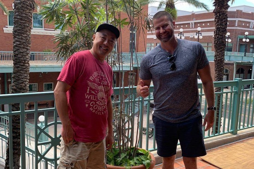 Two people standing on a balcony with palm trees and a building in the background. 3