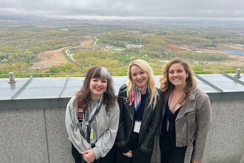 Three individuals standing on a rooftop with a scenic view of a valley and winding roads. 2