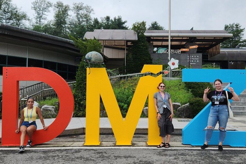 Three people posing in front of large colorful letters spelling 'DMZ'. 4