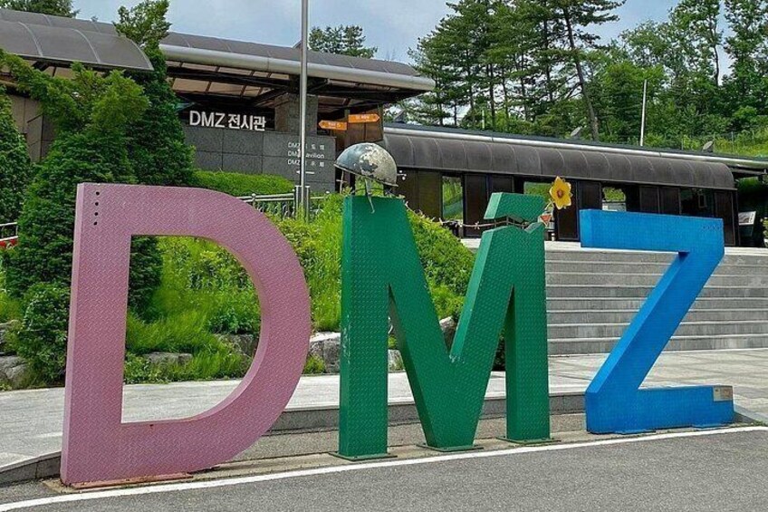 Large colorful letters D, M, and Z, a green trash bin, and a building with signage in the background. 2