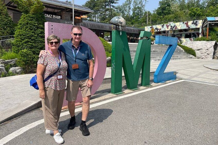 Two people standing in front of a large, colorful 'MZ' sign with a spherical object on top. 3