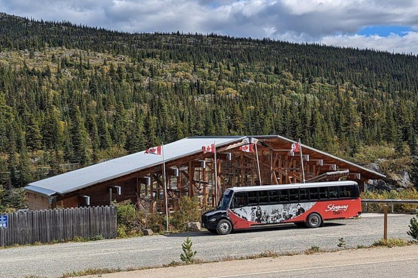 Un autobus est garé devant un bâtiment en bois, orné d’un drapeau canadien, avec en arrière-plan une montagne boisée. 4