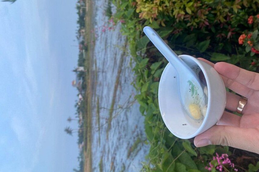 Une main tenant une tasse blanche avec une cuillère, contenant une substance jaune, en extérieur, avec de la verdure et une étendue d'eau en arrière-plan. 2