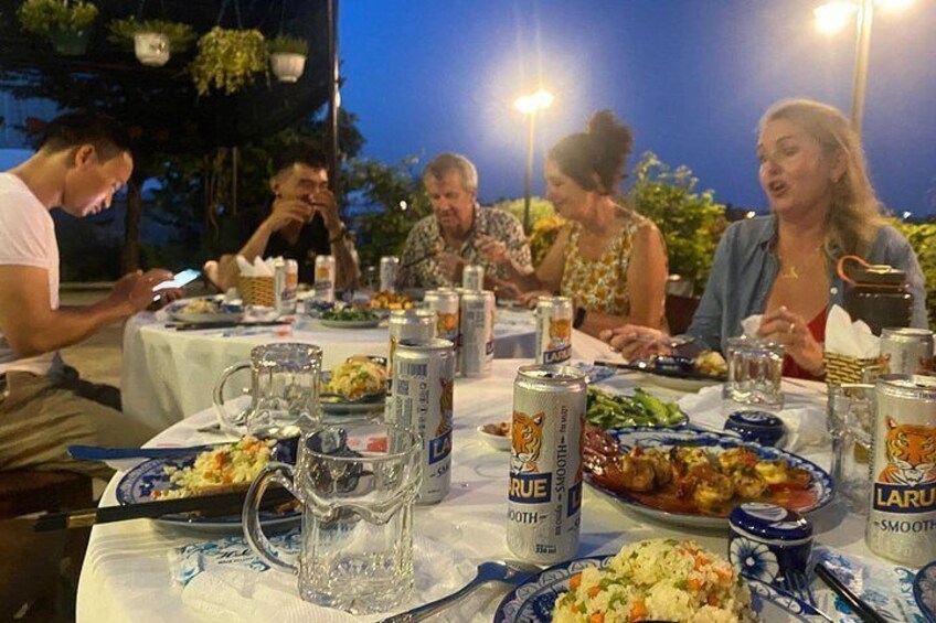 Un gruppo di persone cena all'aperto di sera, a tavoli apparecchiati con piatti, bevande e una caraffa. 2