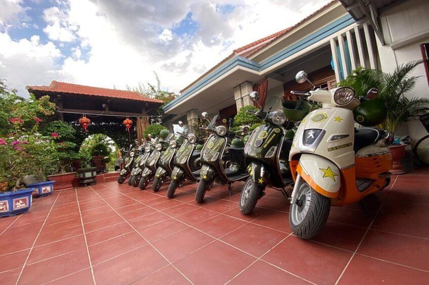 Une rangée de scooters est garée devant un bâtiment dont le sol est recouvert de carreaux et où l’on peut voir des plantes en pot. 5