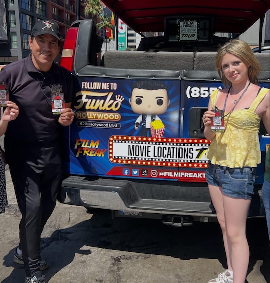 Two people holding VIP passes in front of a vehicle with 'Film Freak Movie Locations' and 'Follow Me to Fungo Hollywood' signage. 2