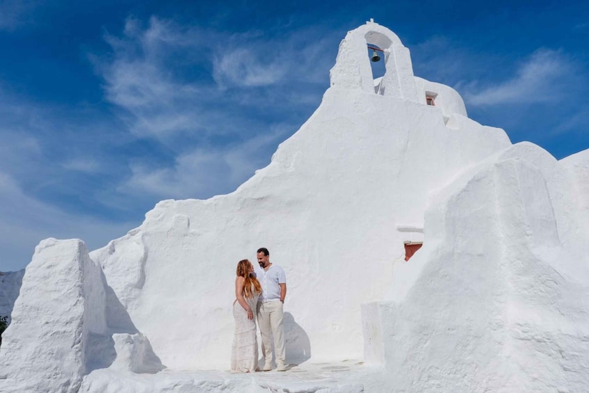 A couple standing in front of a white building with a bell tower, under a clear blue sky. 5