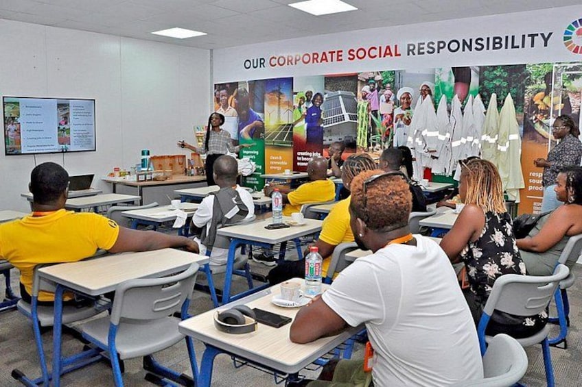 Un'aula scolastica con studenti seduti ai banchi, un insegnante che espone alla lavagna e un poster al muro sulla responsabilità sociale d'impresa. 2
