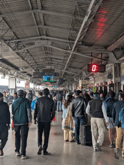 A crowded indoor space with a metal roof, people walking, and a digital display showing '0:'. 2