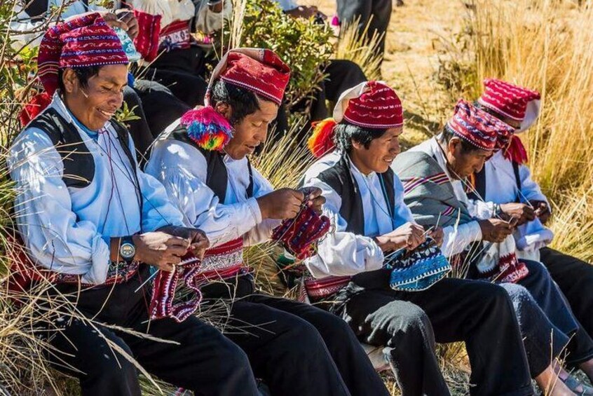 Personas sentadas al aire libre, vestidas con atuendo tradicional, sombreros rojos y camisas blancas, realizando una actividad con artículos de tejido colorido. 2