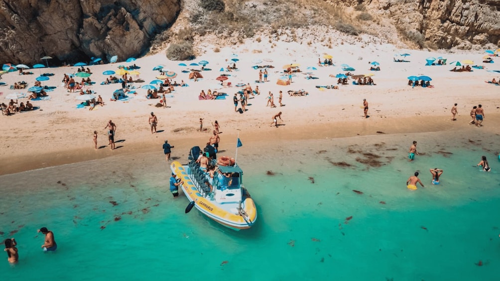 A crowded beach with people sunbathing and swimming, a lifeboat, and clear turquoise water. 1