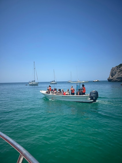 A boat with passengers on a turquoise sea, a rocky coastline, and sailboats in the distance. 2