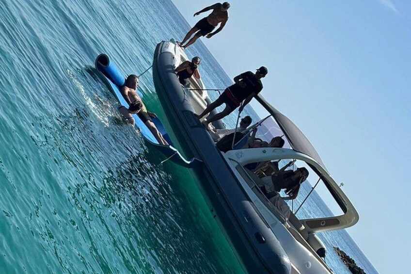 A boat with people on it, a person swimming, and a clear blue sea. 2