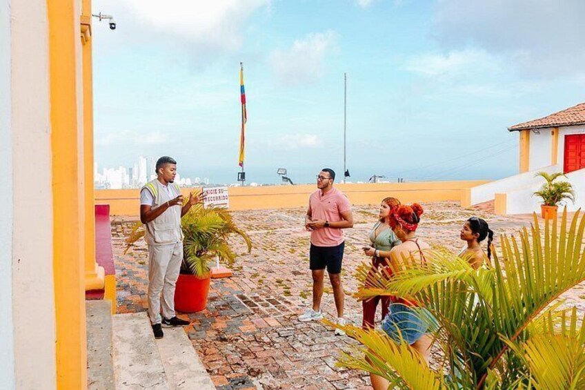 A group of people standing on a rooftop terrace with potted plants, a flagpole, and a city skyline in the background. 5
