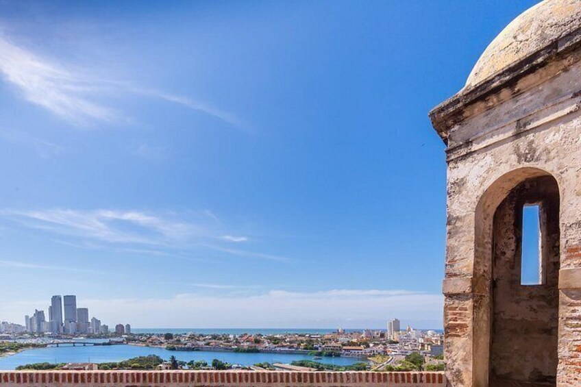A coastal cityscape with a prominent historical stone structure, a clear blue sky, and a distant urban skyline. 3