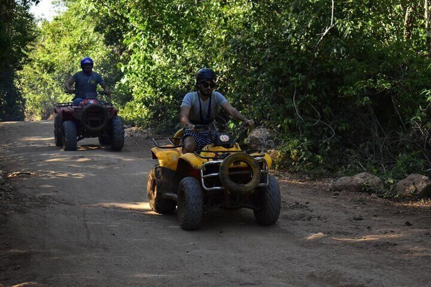 Two people riding ATVs on a dirt road surrounded by trees. 1