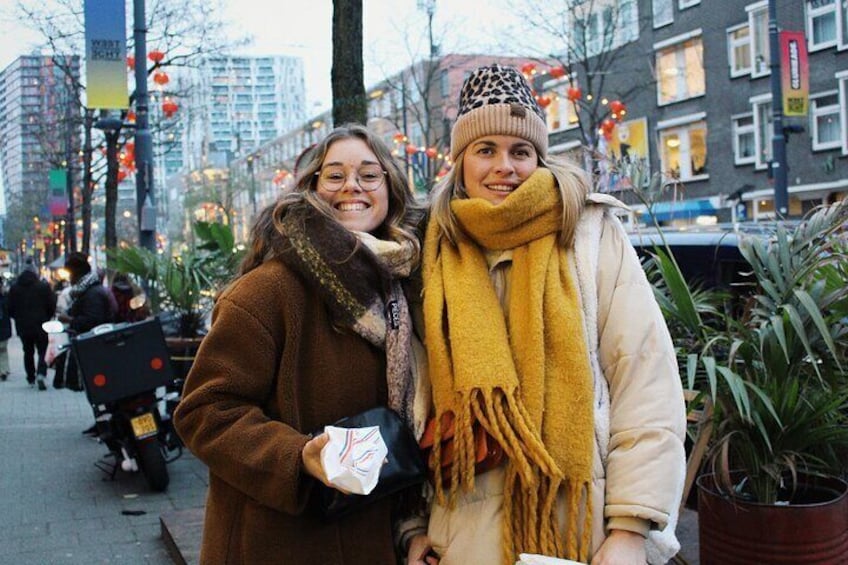 Twee personen staan op een straat met gebouwen en planten op de achtergrond. 5