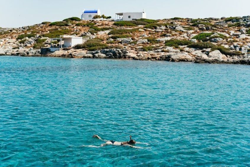 A swimmer in a turquoise sea with a rocky coastline and white buildings in the background. 3