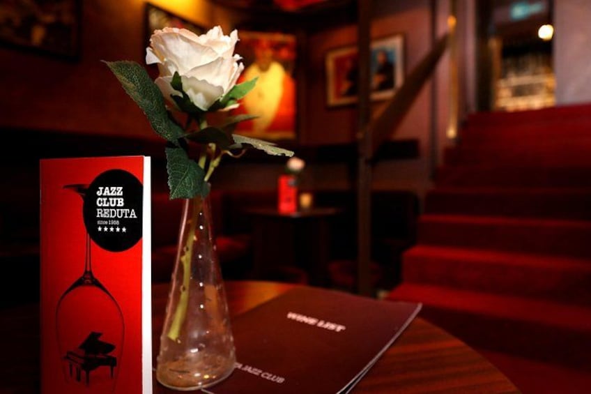 Une boîte rouge portant l’inscription « JAZZ CLUB REDUTA », une rose blanche dans un vase transparent et un menu posé sur une table en bois. 2