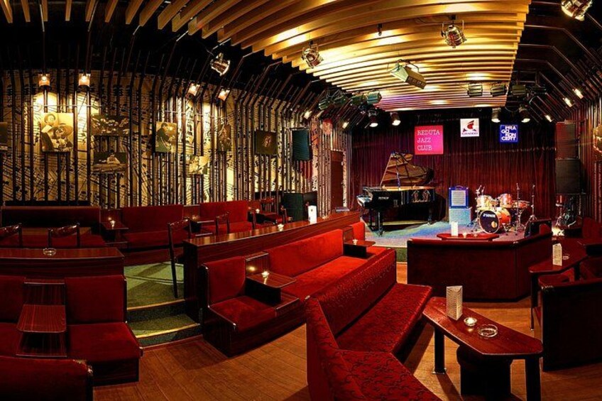 En svagt upplyst lounge med röda sittplatser, en scen med musikinstrument och en skylt med texten ”DENTAL CLUB”. 2