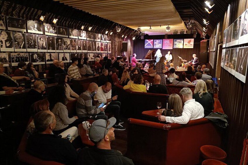 En fullsatt bar med folk sittande vid borden, en scen med uppträdande artister och inramade tavlor på väggarna. 3