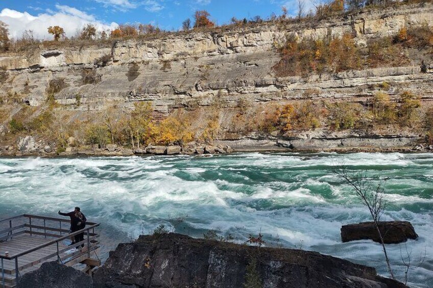 A river with white water rapids, a rocky cliff, and a person standing on a wooden platform. 5