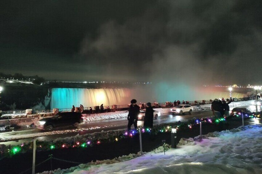 A nighttime scene with a waterfall illuminated by colorful lights, a snow-covered landscape, and a few people standing near the water. 3