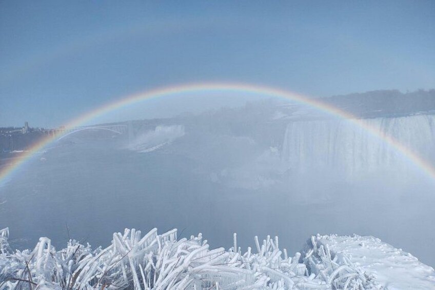 A waterfall with a rainbow, snow-covered ground, and a clear sky. 3