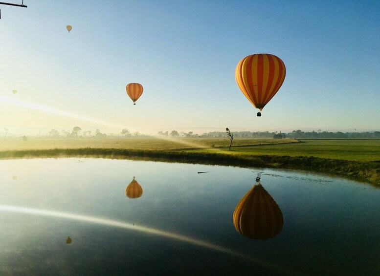 Hot air balloons over a calm water body with reflections, surrounded by open fields and trees. 2
