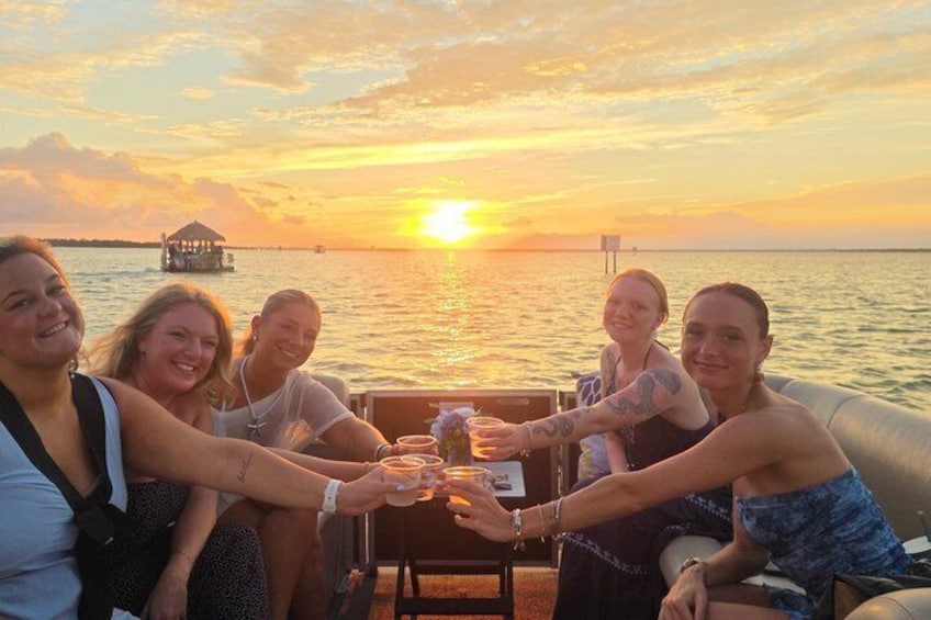 A group of people toasting with drinks on a boat at sunset with a pier and water in the background. 1